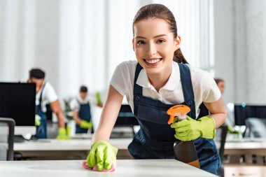 happy cleaner looking at camera while cleaning office desk with rag clipart