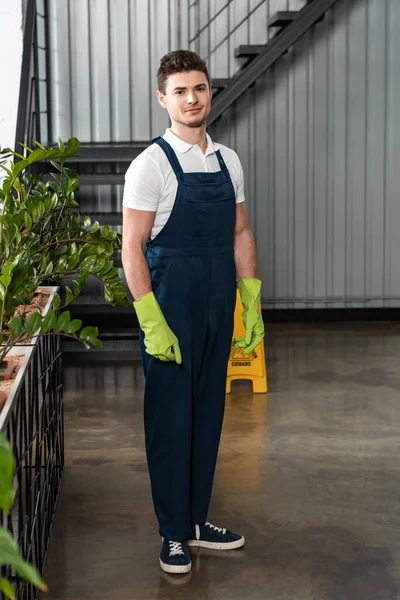 smiling young cleaner in overalls and rubber gloves looking at camera