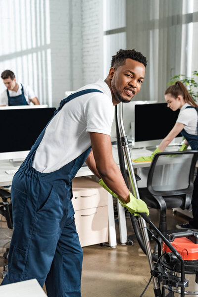 handsome african american cleaner moving vacuum cleaner while smiling at camera