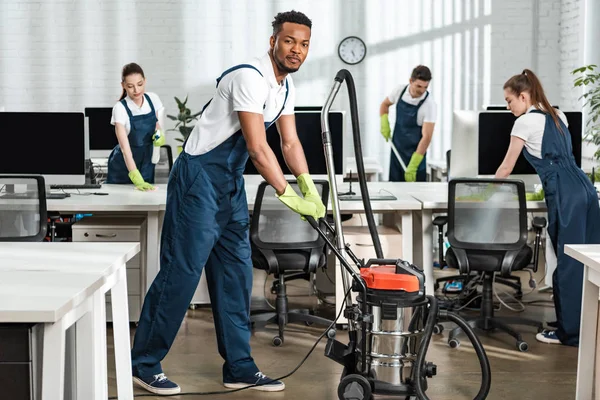 Tersenyum African American Cleaner Bergerak Vacuum Cleaner Dekat Tim Rekan — Stok Foto