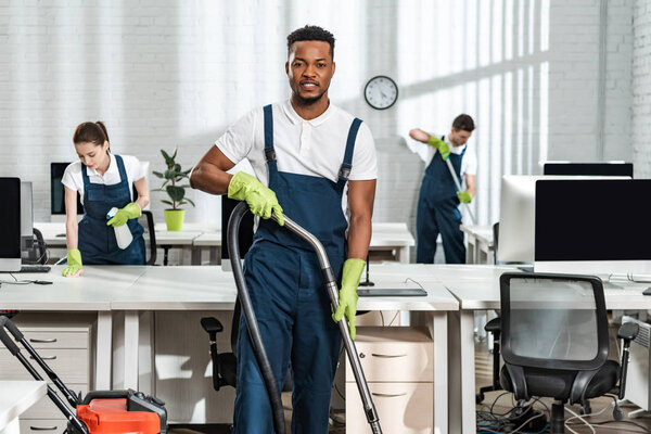 handsome african american cleaner looking at camera while standing with vacuum cleaner near colleagues