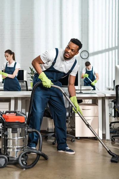 Sorrindo Afro Americano Limpador Aspirando Chão Enquanto Olha Para Longe — Fotografia de Stock