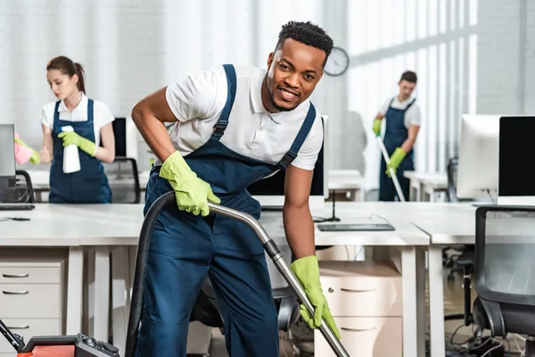 Fröhliche Afrikanisch Amerikanische Putzfrau Staubsaugt Boden Während Sie Die Kamera — Stockfoto