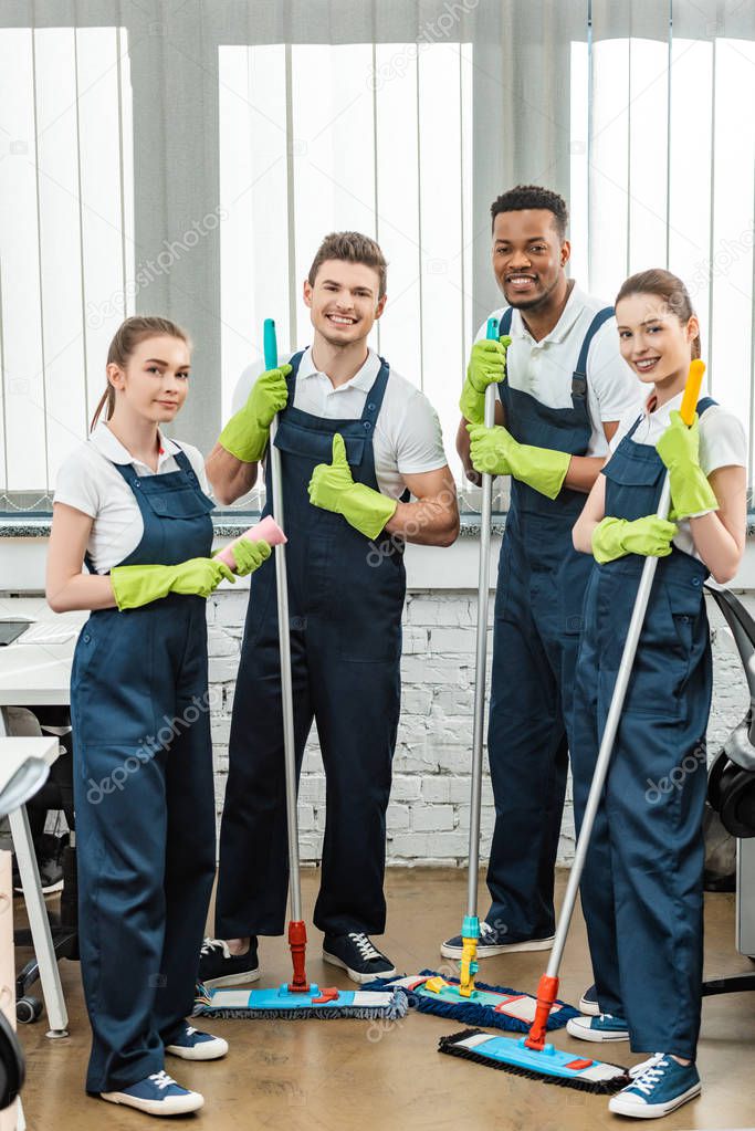 happy multicultural team of cleaners looking at camera and showing thumbs up while holding mops