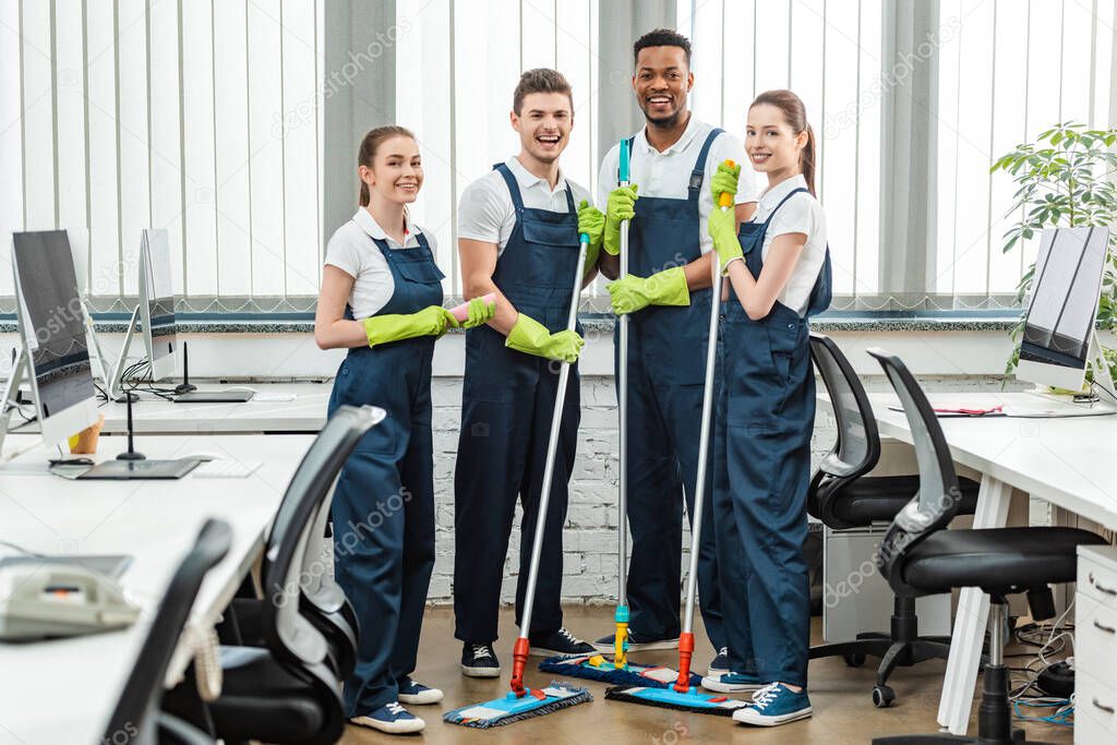 cheerful multicultural team of cleaners looking at camera while standing with mops in office