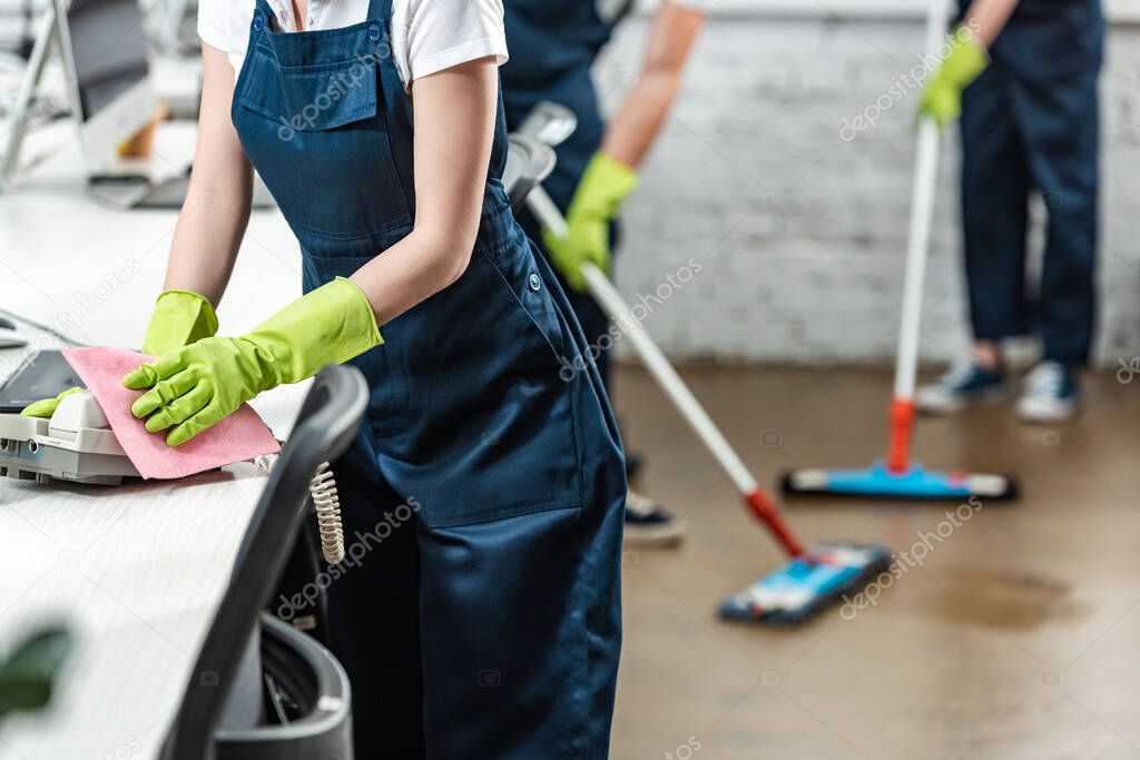 cropped view of cleaner wiping phone while colleagues washing floor with mops