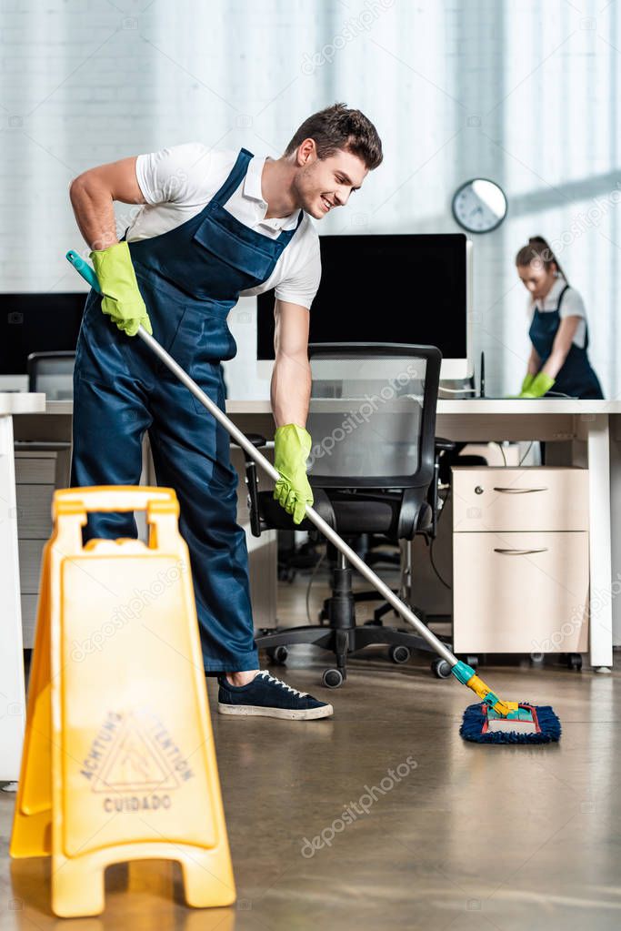 smiling cleaner washing floor with mop near colleague cleaning desk