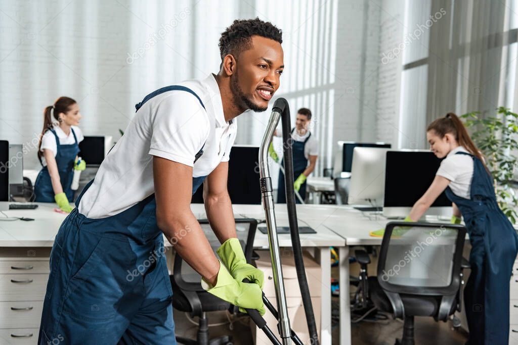 smiling african american cleaner moving vacuum cleaner near team of colleagues
