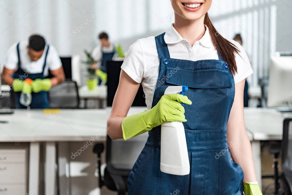 cropped view of smiling cleaner in overalls holding detergent spray