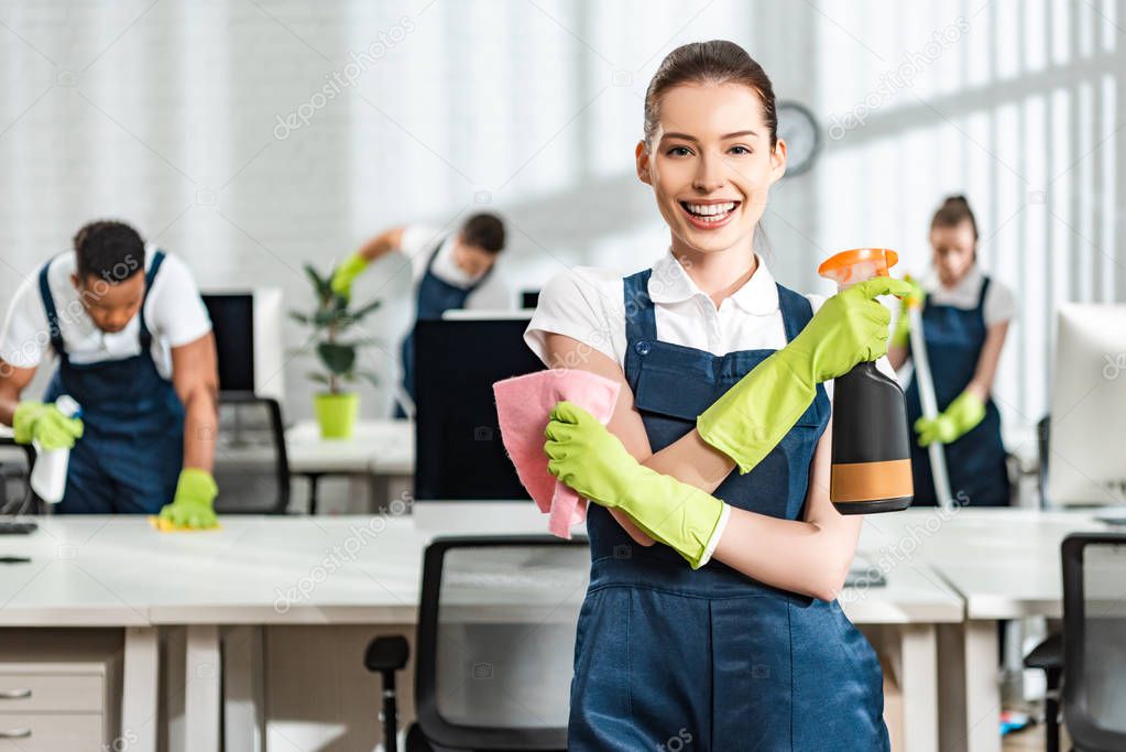 happy cleaner with spray and rag smiling at camera while standing near multicultural colleagues