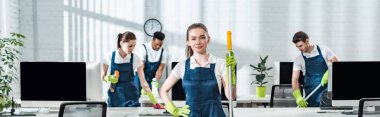 panoramic shot of smiling cleaner standing with hand on hip near multicultural colleagues clipart