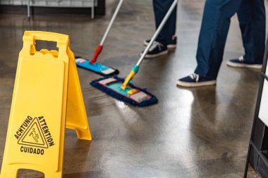 partial view of two cleaners washing floor with mops near wet floor caution sign clipart
