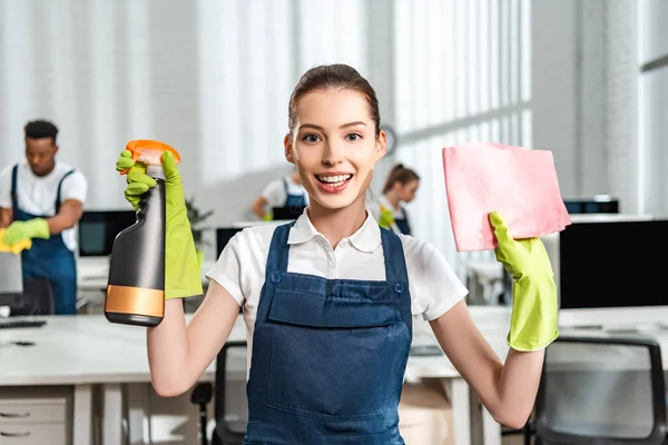 happy cleaner in overalls holding spray bottle and rag while looking at camera