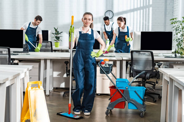 attractive cleaner holding mop while standing with hand on hip near multicultural colleagues