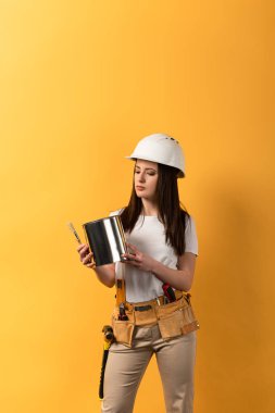 serious handywoman holding paint can and paint brush on yellow background  clipart