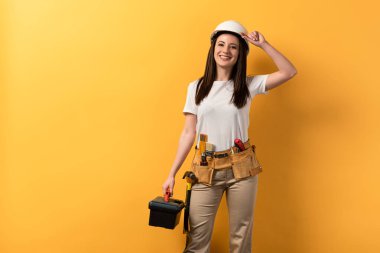 smiling handywoman in helmet holding toolbox on yellow background  clipart