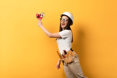 shocked handywoman holding tape dispenser on yellow background with copy space  clipart