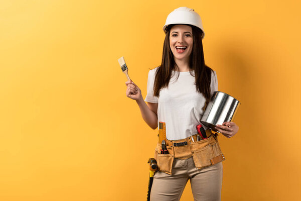smiling handywoman holding paint brush and paint can on yellow background 