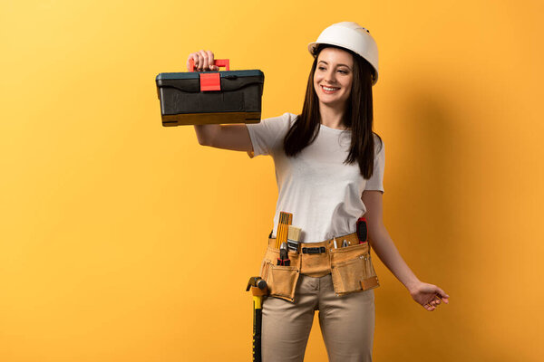 smiling handywoman in helmet holding toolbox on yellow background 