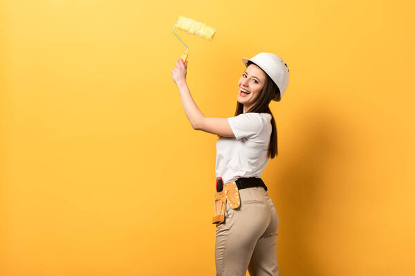 smiling handywoman holding paint roller on yellow background with copy space