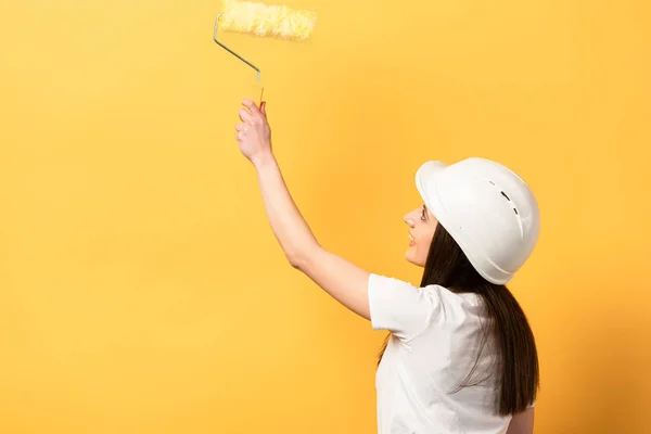 smiling handywoman holding paint roller on yellow background