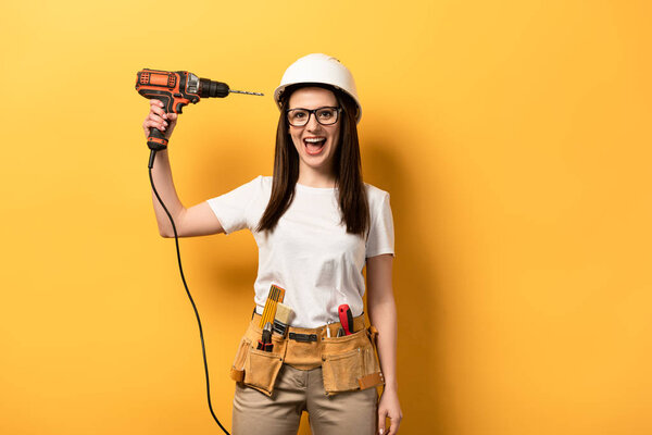 smiling handywoman holding drill and looking at camera on yellow background 