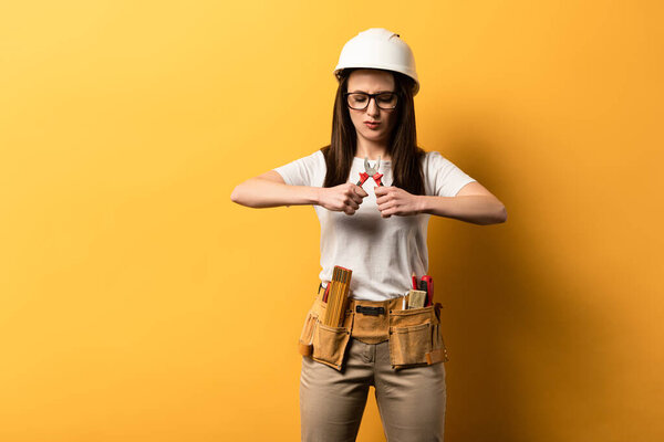 concentrated handywoman in helmet holding pliers on yellow background 