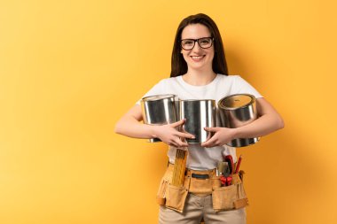 smiling repairwoman holding paint cans and looking at camera on yellow background  clipart