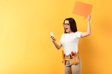 Konuşma baloncuğu tutan ve sarı arka planda akıllı telefon kullanan şok olmuş tamirci kadın. 