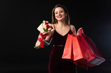 Elinde alışveriş poşetleri ve siyah arka planda buket tutan gülümseyen kadın. 