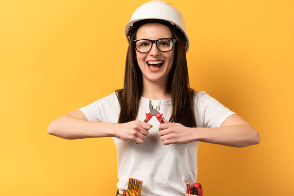 smiling handywoman holding pliers and looking at camera on yellow background 