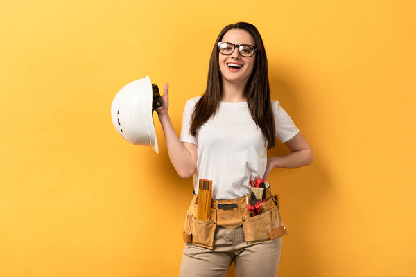 smiling handywoman looking at camera and holding helmet on yellow background 