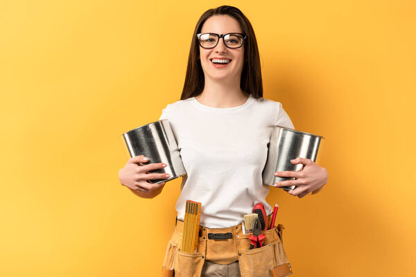 smiling handywoman holding paint cans and looking at camera on yellow background 