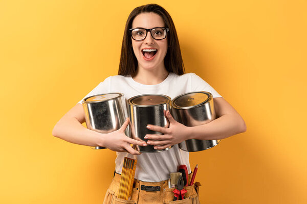 shocked repairwoman holding paint cans and looking at camera on yellow background 