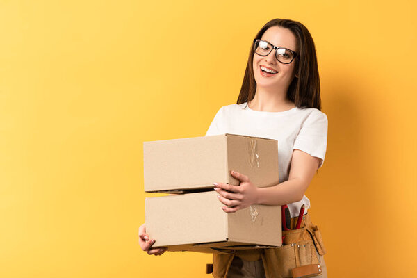 smiling repairwoman holding boxes and looking at camera on yellow background 