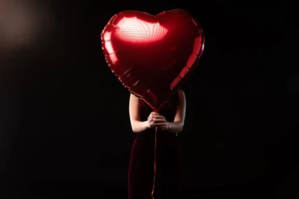 woman in dress obscuring face with heart-shaped balloon in 14 february on black background