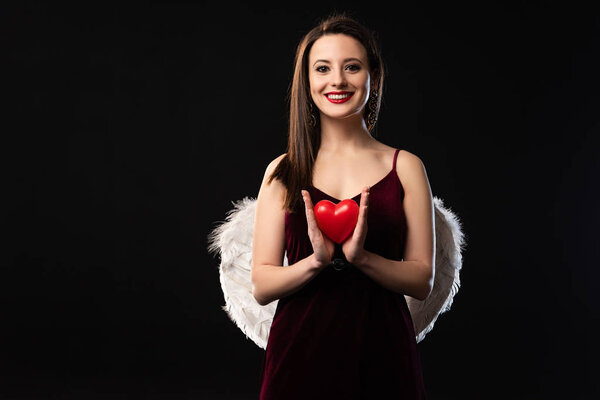 smiling woman in dress with wings holding heart-shaped model in 14 february isolated on black