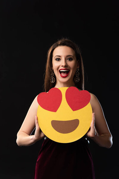 KYIV, UKRAINE - DECEMBER 16, 2019: shocked woman in dress holding smiley isolated on black 