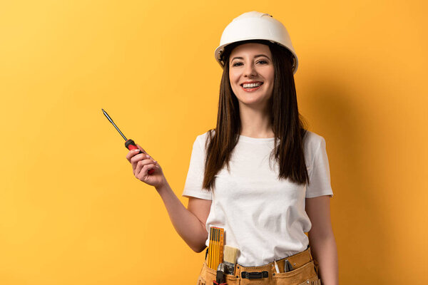 smiling handywoman in helmet holding screwdriver on yellow background 