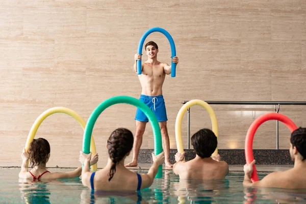 Group of young people exercising with pool noodles and handsome trainer in swimming pool