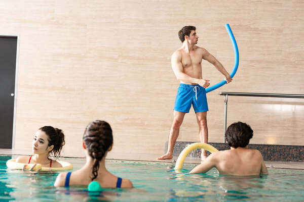 Selective focus of trainer holding pool noodle while exercising with people in swimming pool