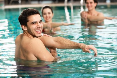 Yüzme havuzunda genç insanlarla egzersiz yaparken kameraya bakan gülen antrenör.