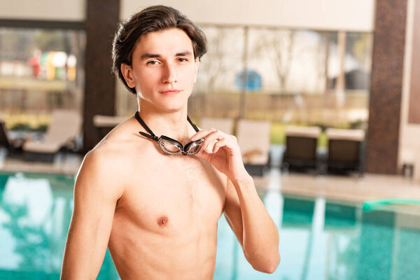 Handsome man holding swimming googles and looking at camera near swimming pool