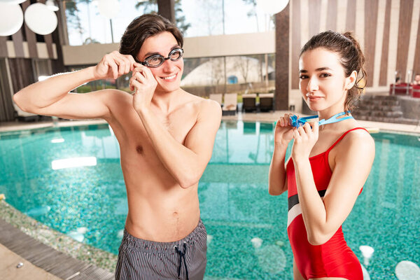 Smiling man and woman holding swimming googles near swimming pool