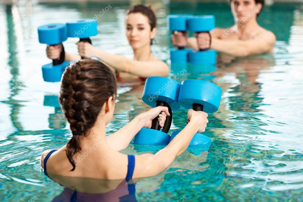 Selective focus of trainer holding barbells while exercising water aerobics with man and woman in swimming pool