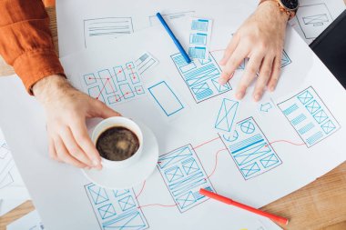 Cropped view of developer working with ux website templates and holding coffee cup on table clipart