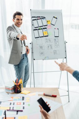 Selective focus of smiling ux designer near whiteboard with templates pointing on colleague with smartphone in office  clipart