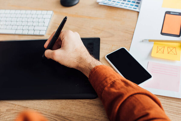 Cropped view of developer using graphics tablet near smartphone and templates of mobile ux design on table