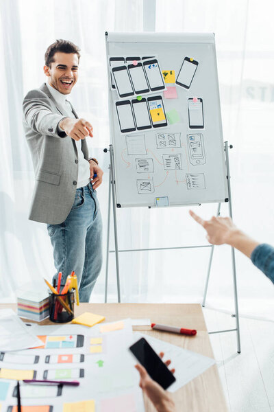 Selective focus of smiling ux designer near whiteboard with templates pointing on colleague with smartphone in office 