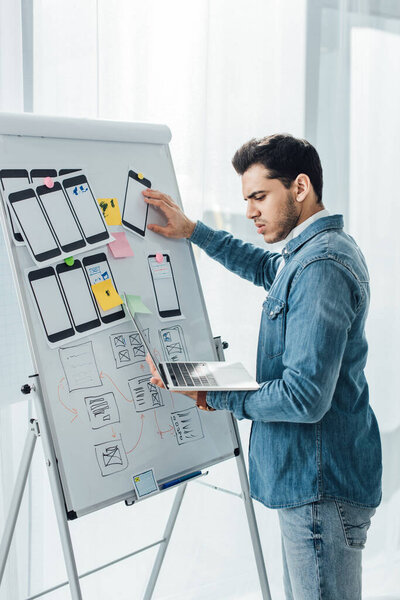 Side view of ux designer using laptop near layouts of mobile frameworks on whiteboard in office 